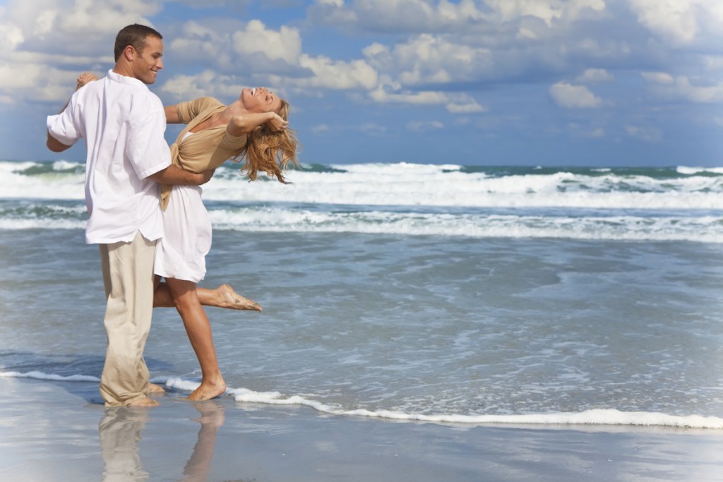 A young man and woman having fun dancing as a romantic couple on a beach with a bright blue sky